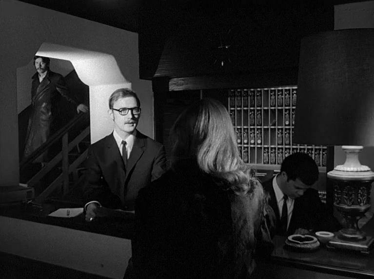 Gods Of The Plague (1970) movie still: medium — A dark hotel lobby features a reception desk with a grid of room keys. A clerk addresses a customer…