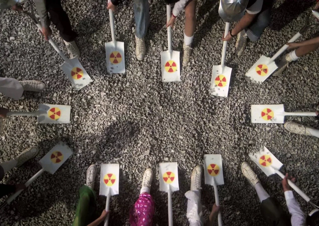 Human Highway (1982) movie still: wide — People stand in a circle on a gravel surface holding shovels inward. Each shovel blade displays a red and…