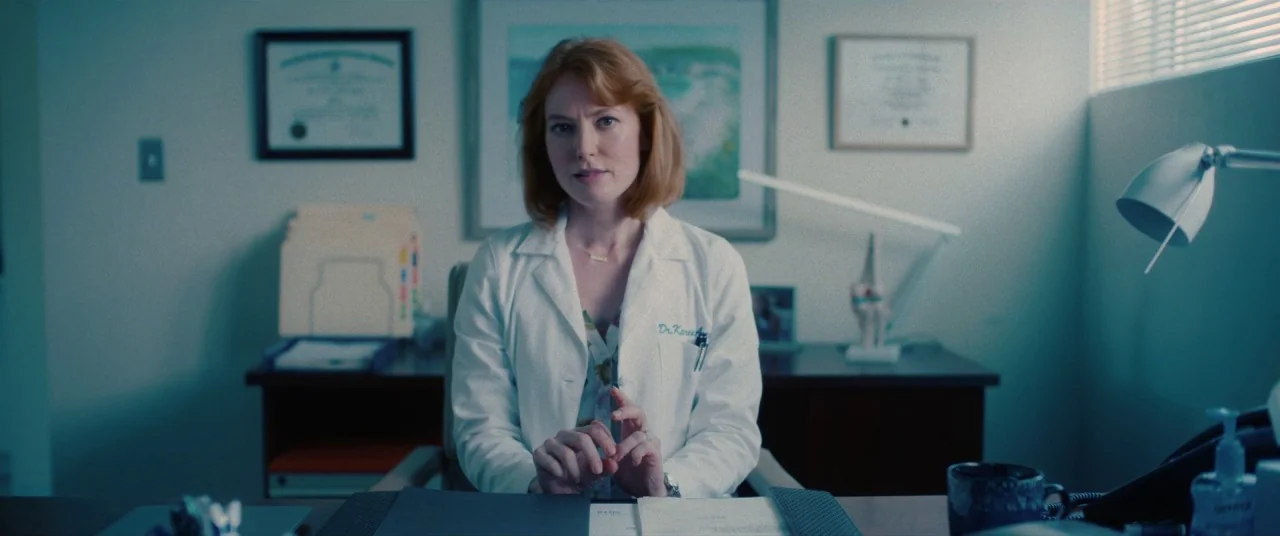 I Care A Lot (2020) movie still: medium — A doctor's office with framed diplomas on the wall behind the subject's desk. The subject sits centered at…