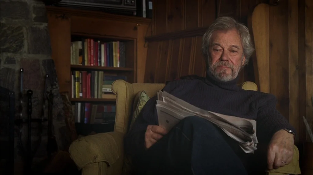 Away From Her (2006) movie still: medium — A cozy room features wood paneled walls, a stone fireplace, and a full bookshelf. An older man sits in a…