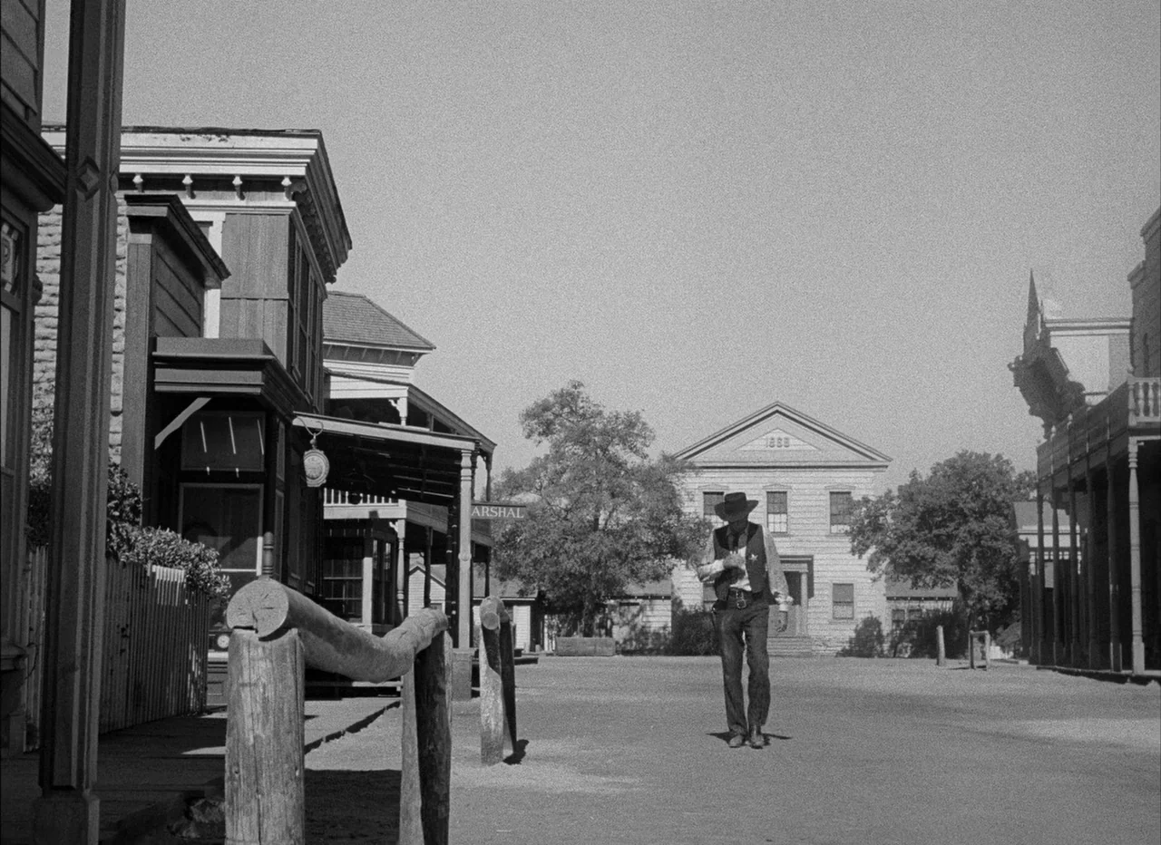 High Noon (1952) movie still: fullbody — A dusty, empty main street in an 1880s wooden frontier town. A lone, armed man walks slowly toward the…