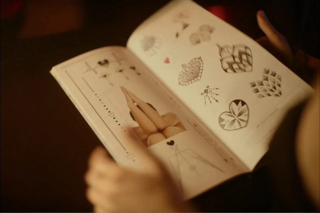Cross the Line (2020) movie still: closeup — An open book with pages displaying various black ink tattoo sketches and a small photograph. Hands hold…