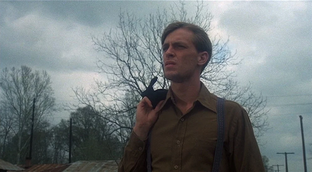 Thieves Like Us (1974) movie still: medium — A man stands in an open area under a grey, overcast sky with leafless trees. He looks toward the…