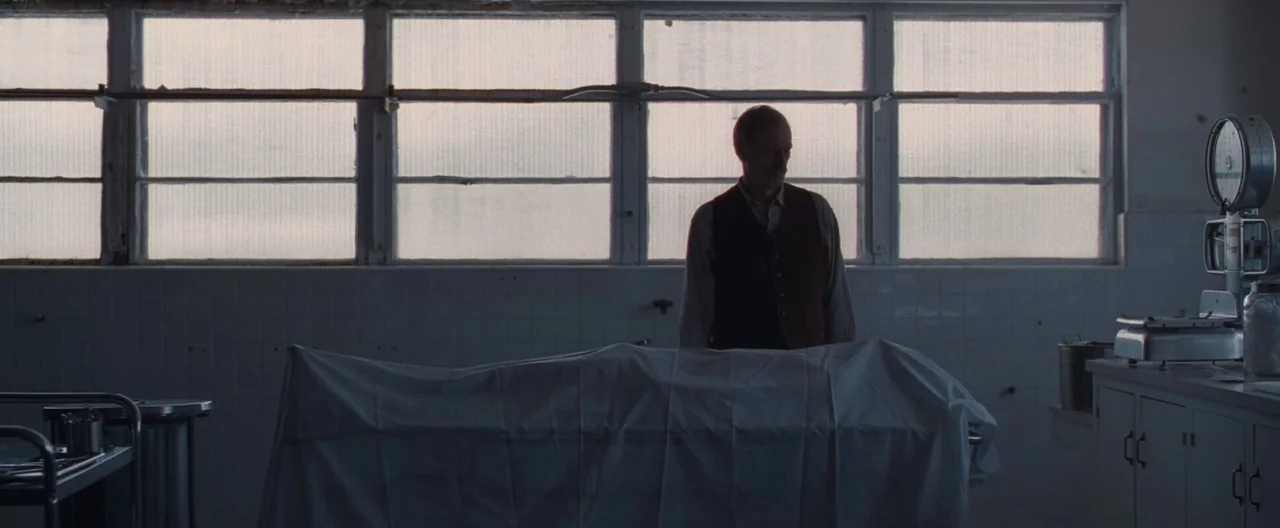 Ain’t Them Bodies Saints (2013) movie still: medium — A window-lined morgue features tiled walls and metal work surfaces. An elderly man in a vest…