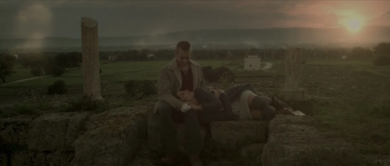 Spring (2014) movie still: wide — Ancient stone ruins with scattered pillars overlook a flat agricultural valley at sunset. A man and a woman sit…