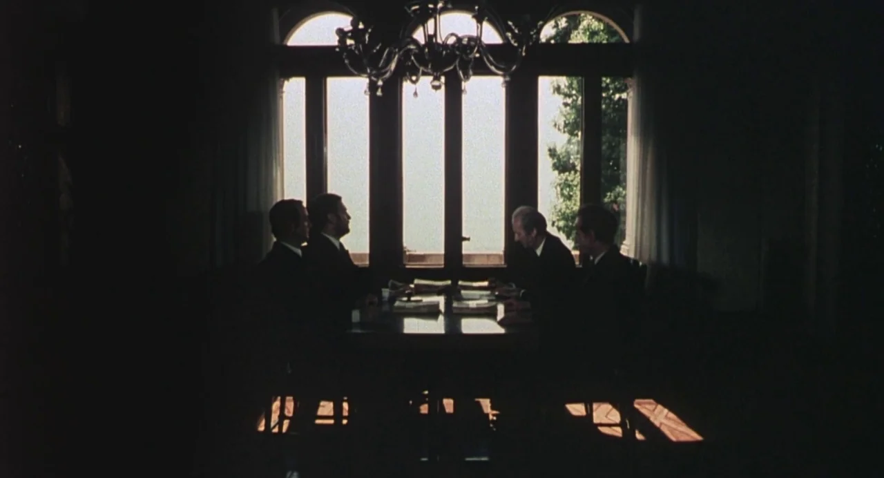 Salò, or the 120 Days of Sodom (1975) movie still: wide — A dark room with four men sitting around a table against a window. They are holding papers…