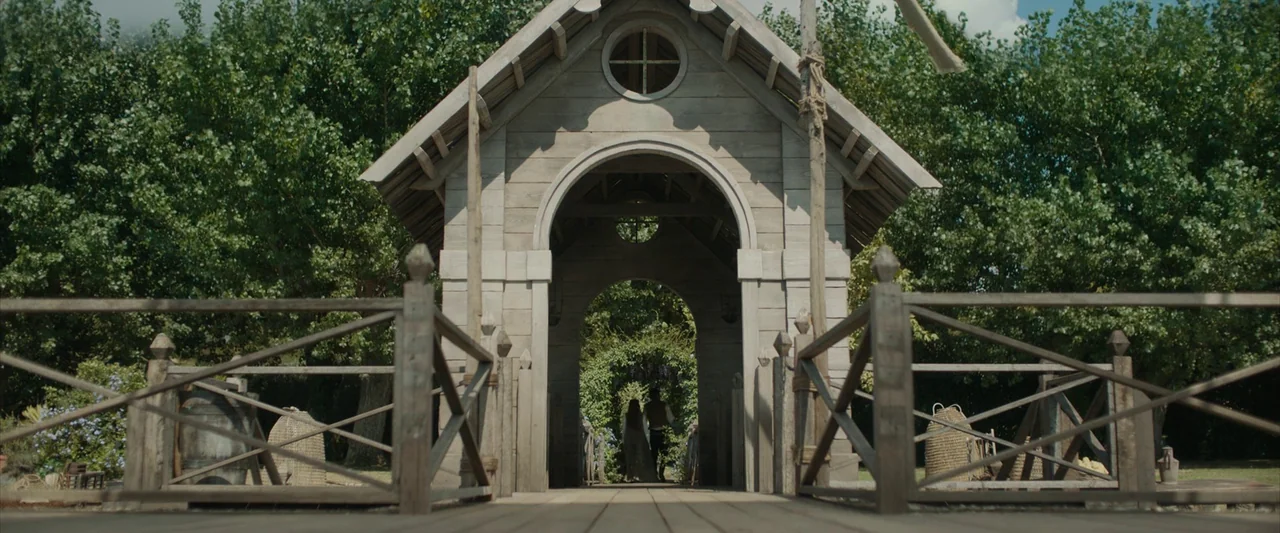 Rosaline (2022) movie still: wide — A wooden bridge with a gabled roof spans a passage between dense green trees. A man and woman in historical…