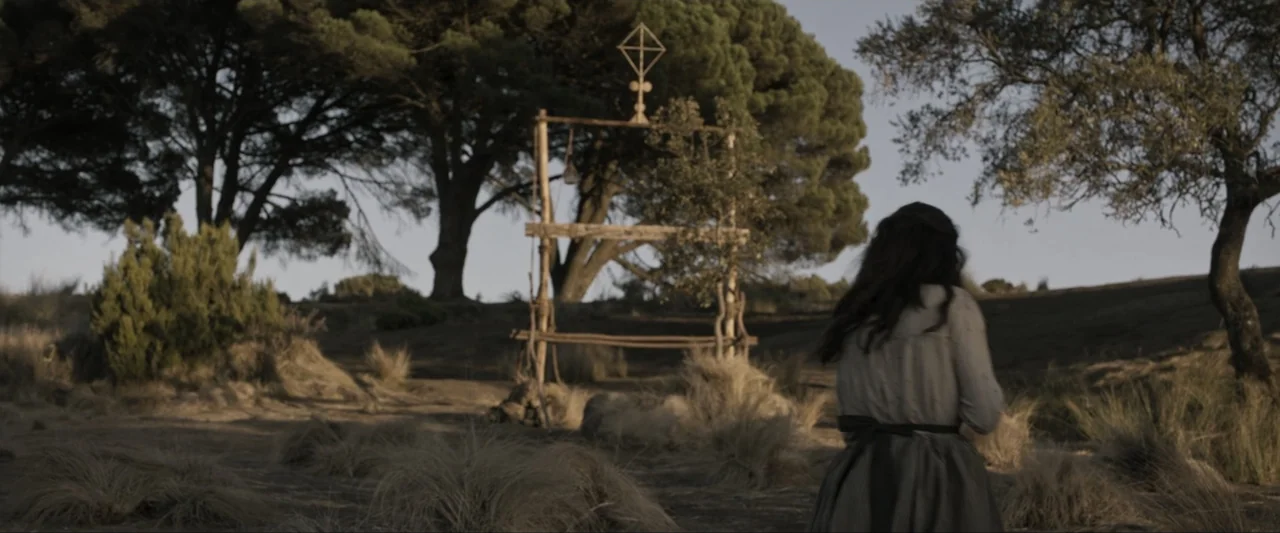Fatima (2020) movie still: medium — An open dry field contains a solitary wooden frame structure with a geometric cross. A woman in a…