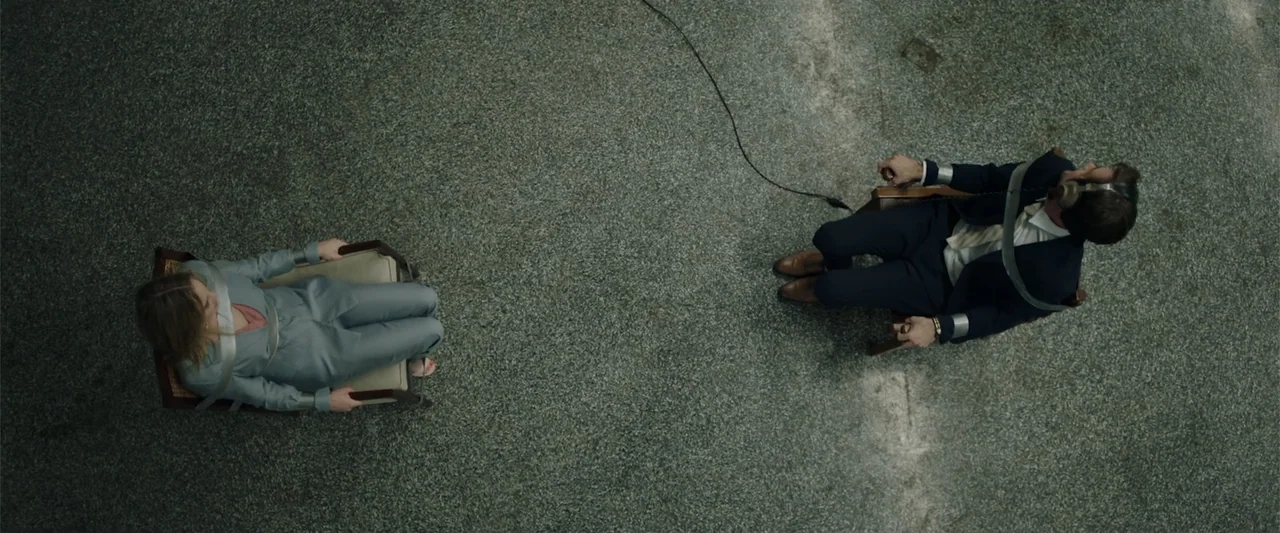Playing God (2021) movie still: wide — A sterile, minimalist room features two subjects secured in wooden chairs on concrete floor. The subjects sit…