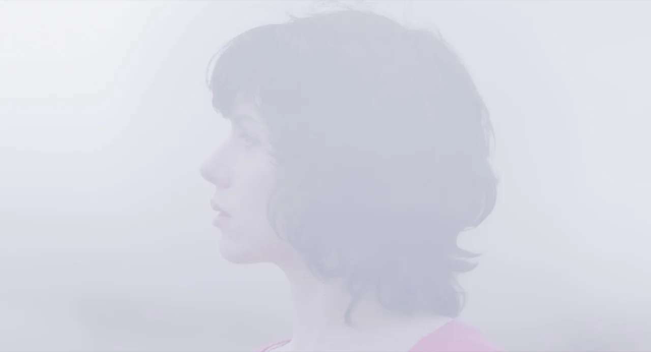 Under The Skin (2013) movie still: closeup — A figure stands in a thick, pervasive fog obscuring much of the surroundings. The person is captured in…