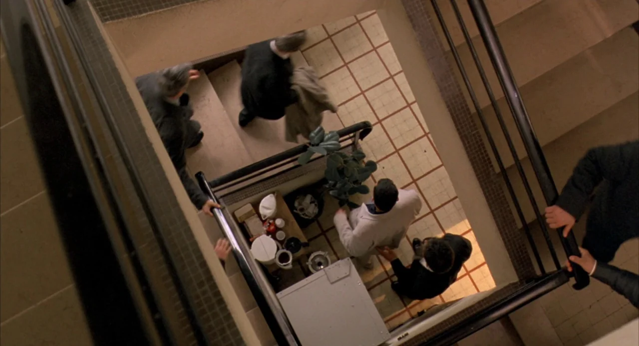 The Good Thief (2002) movie still: wide — An interior concrete stairwell with off-white floor tiles and a dark metal railing system. Several men in…