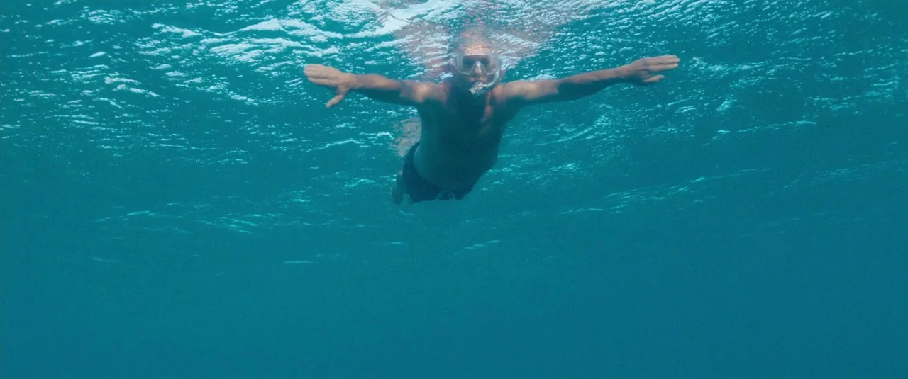 A Hologram for the King movie still: wide — A man floats on the water surface in a clear blue open ocean. The subject wears snorkeling gear and…