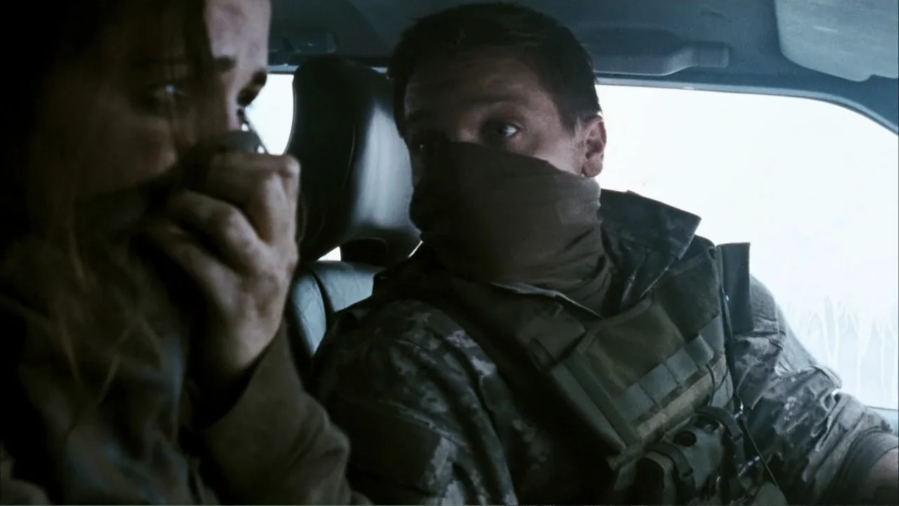 28 Weeks Later (2007) movie still: medium — The interior of a vehicle shows two people sitting in the front row. A man in tactical gear looks toward…