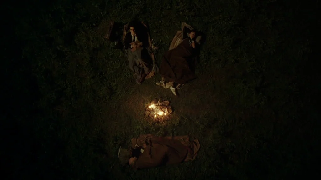 Everything Is Illuminated (2005) movie still: wide — A grassy clearing at night contains three people sleeping around a small, burning campfire. The…