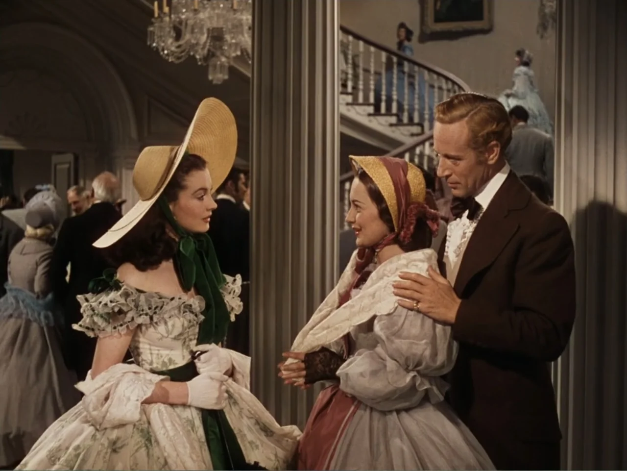 Gone with the Wind (1939) movie still: medium — A crowded interior ballroom features a grand staircase and ornate architectural columns in the…