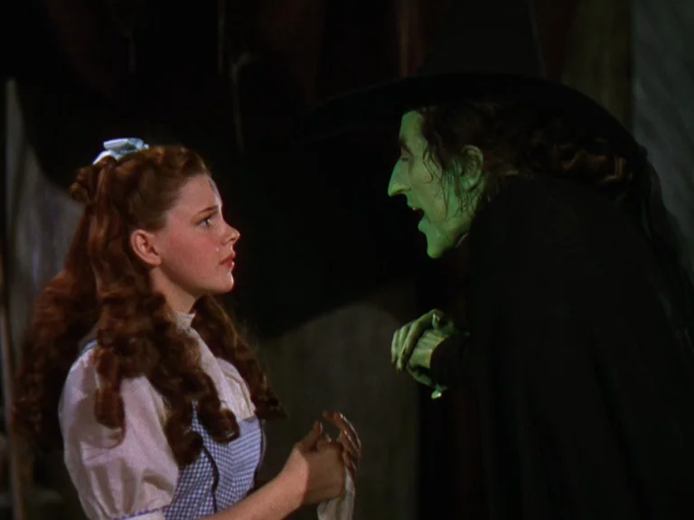The Wizard Of Oz (1939) movie still: medium — An interior stone setting with low light and dark, indistinct walls behind the subjects. A girl faces…