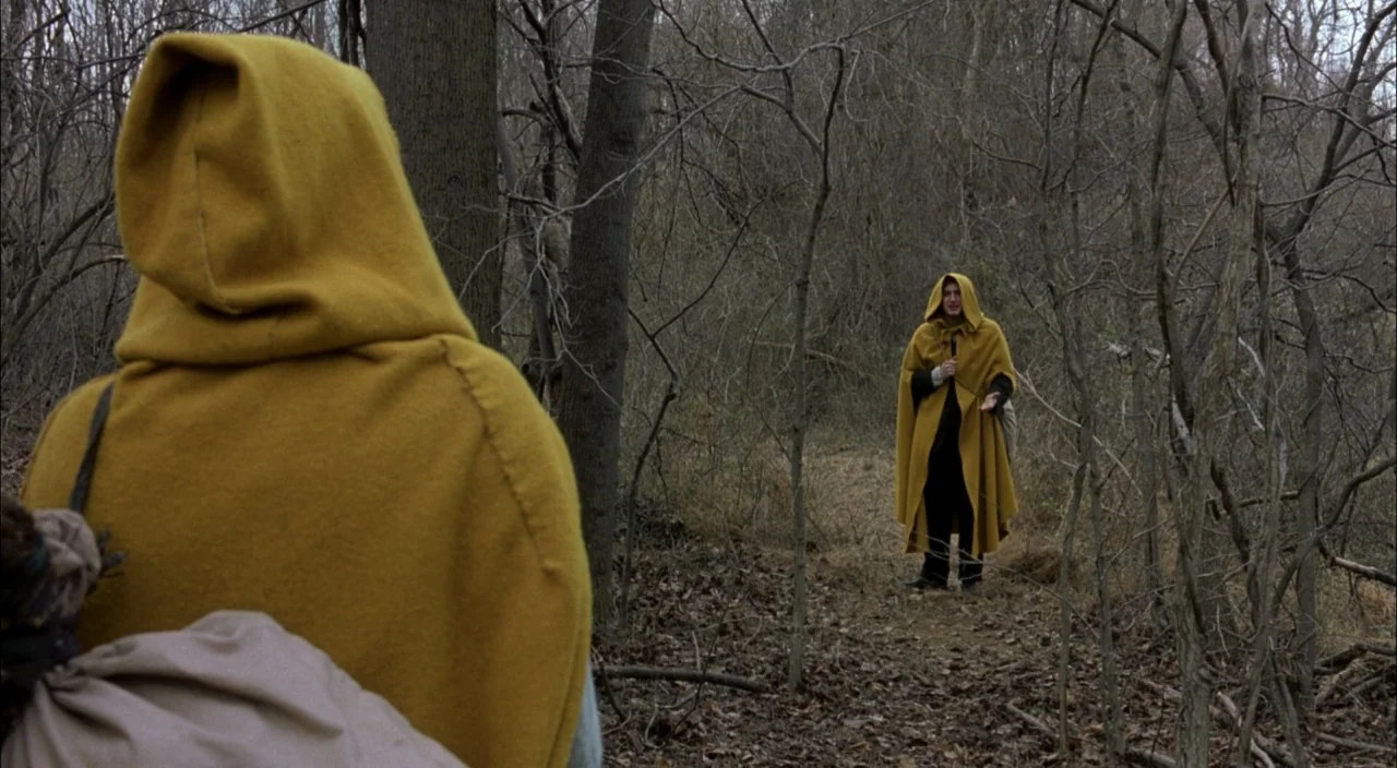 The Village (2004) movie still: medium — A clearing within a dense wood filled with leafless trees and dry brown leaves. A man wearing a yellow…