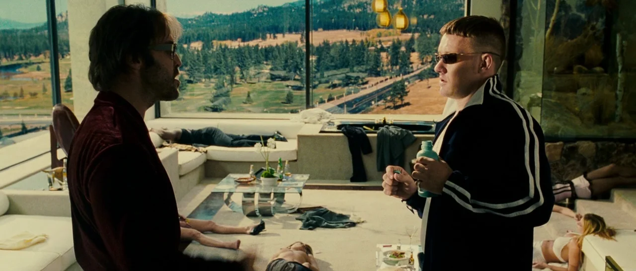 Slice (2018) movie still: medium — A spacious, modern penthouse with floor-to-ceiling windows overlooking a distant pine forest. Several people are…