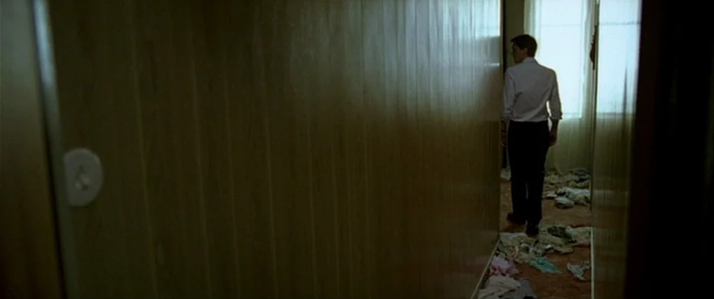 Next Door (2005) movie still: fullbody — A narrow residential hallway features wooden paneling on the walls and cluttered flooring. A man stands at…