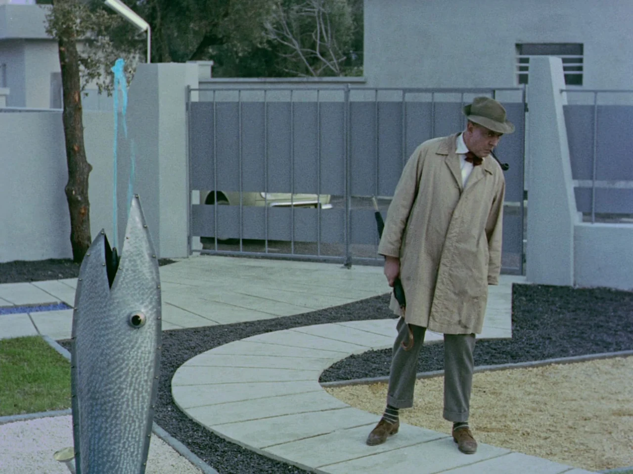 Mon Oncle (1958) movie still: fullbody — A modern suburban home exterior features a stone pathway and minimalist landscaping. A man in a trench coat…