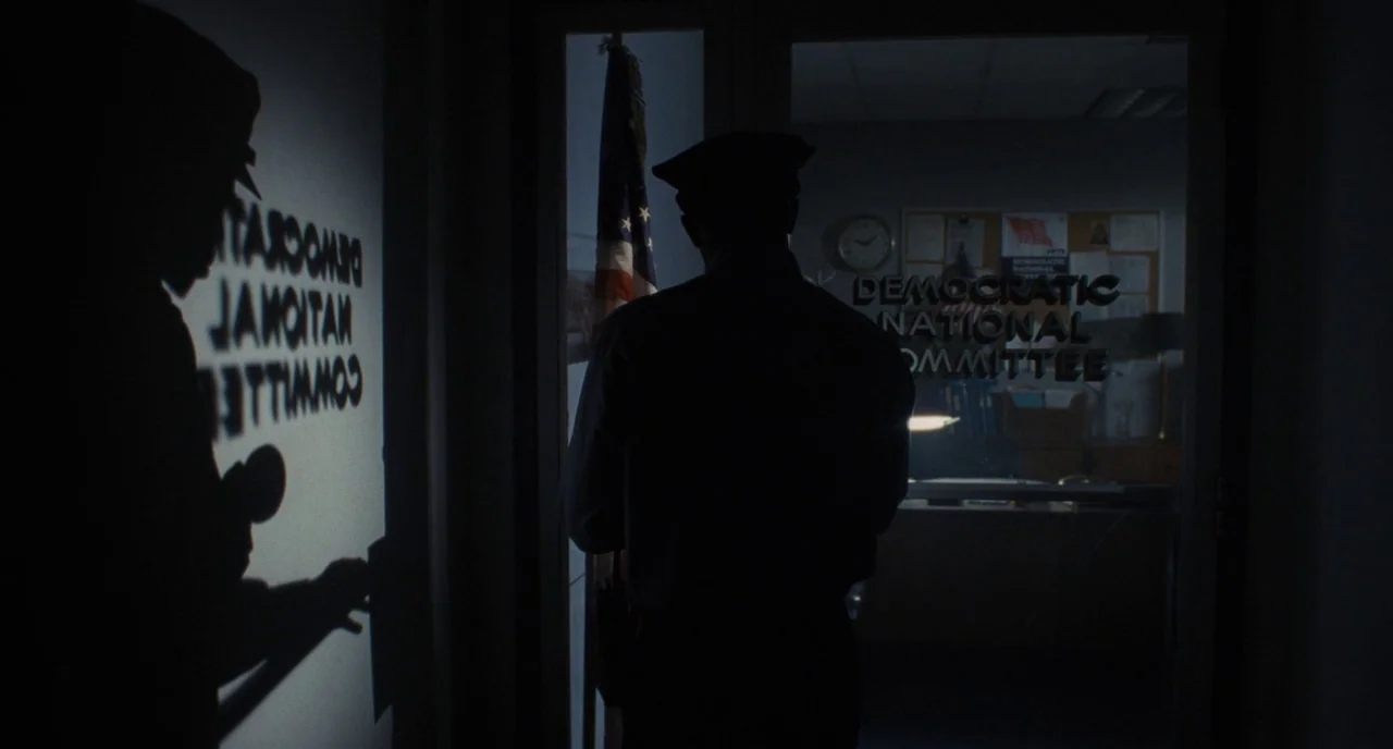 The Post (2017) movie still: medium — A dark hallway leads to an office with a sign reading Democratic National Committee. A guard stands in the…