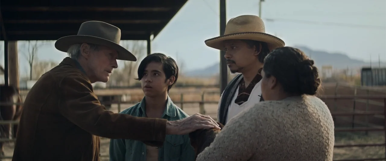 Cry Macho (2021) movie still: medium — An outdoor paddock with a wooden fence and distant hills under soft sunlight. Four people stand together…