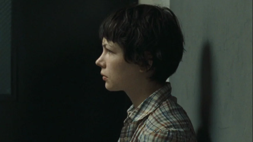 Wendy and Lucy (2008) movie still: medium — A dimly lit indoor space features a wall surface on one side. A woman with short hair stands against the…