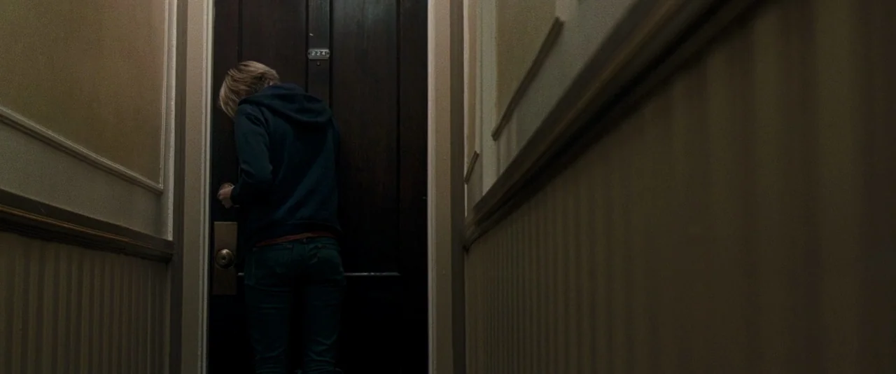 The Innkeepers (2011) movie still: medium — A narrow residential hallway features vertically striped wallpaper and recessed dark wood trim. A woman…