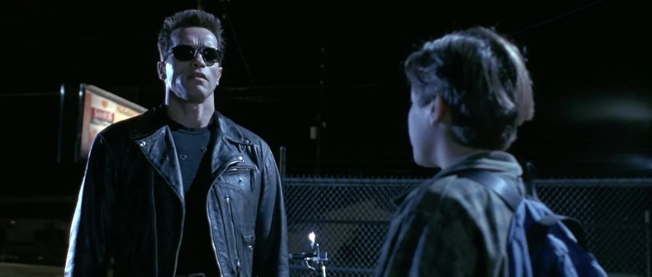 Terminator 2: Judgement Day (1991) movie still: medium — A man in a leather jacket stands before a teenage boy in a parking lot. A chain-link fence…