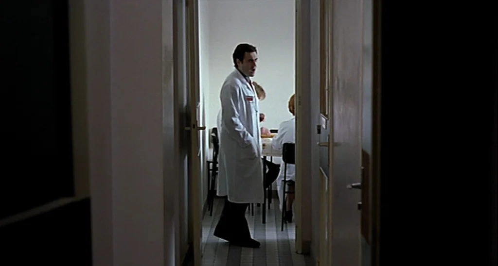 Trouble Every Day (2001) movie still: medium — A spare, clinical room features white walls, a tiled floor, and several simple chairs. A man in a lab…