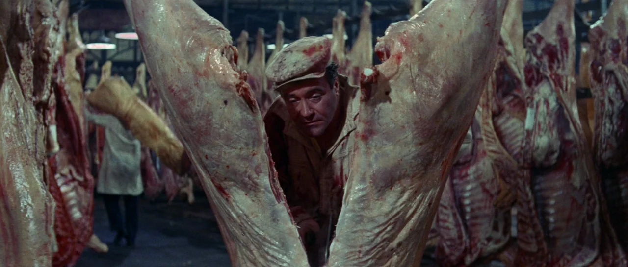 Irma La Douce (1963) movie still: medium — A meat packing plant is filled with rows of hanging slaughtered animal carcasses. A man wearing a work…