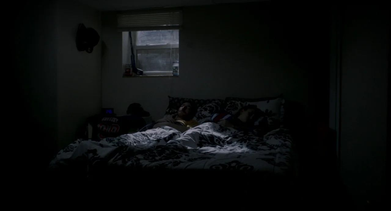 Happer’s Comet (2022) movie still: wide — A dimly lit room features a bed positioned beneath a single small window. Two people sleep under a…