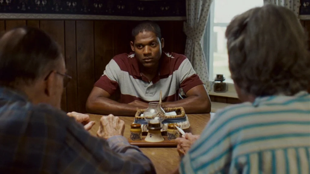 Sugar (2008) movie still: medium — A dining room features wood-paneled walls and wallpaper with a small rooster pattern border. A young man sits at…