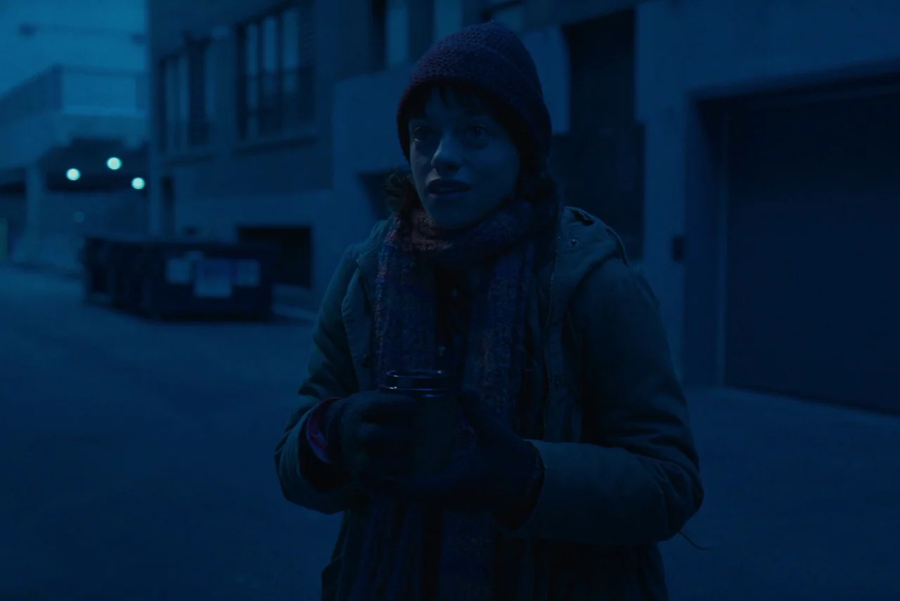 Red Rooms (2023) movie still: medium — A dark city street at night is illuminated by a cold blue color grade. A person in winter gear holds a…