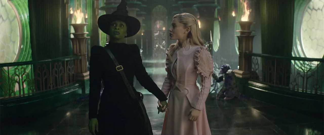 Wicked (2024) movie still: medium — An ornate emerald green hallway features tall arched windows and glowing torch wall sconces. Two women holding…