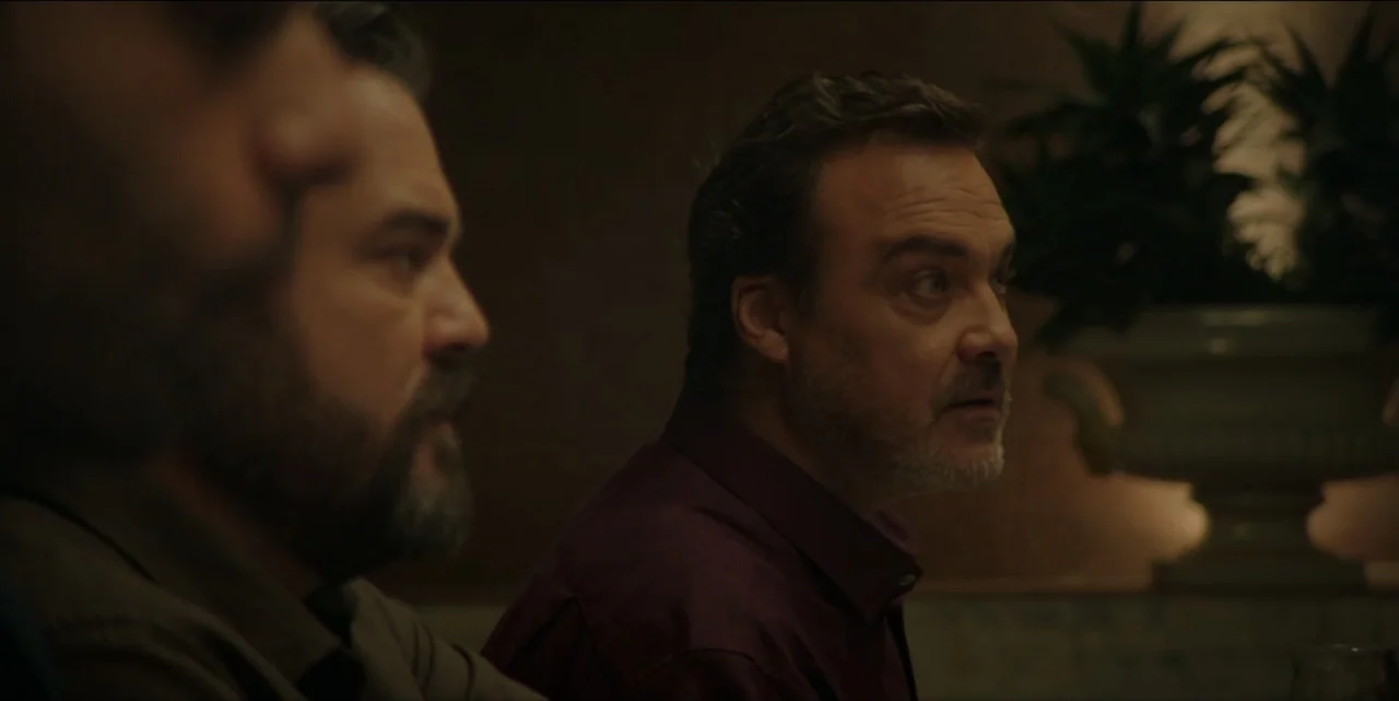 The Hunted (2024) movie still: medium — A dimly lit indoor dining area features dark walls and a potted plant backdrop. Two middle-aged men sit side…