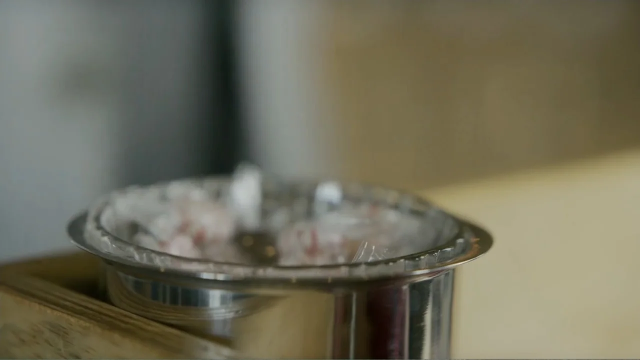 Lecciones para canallas (2022) movie still: closeup — A metal container with a plastic-lined rim rests on a light-colored wooden surface. The…