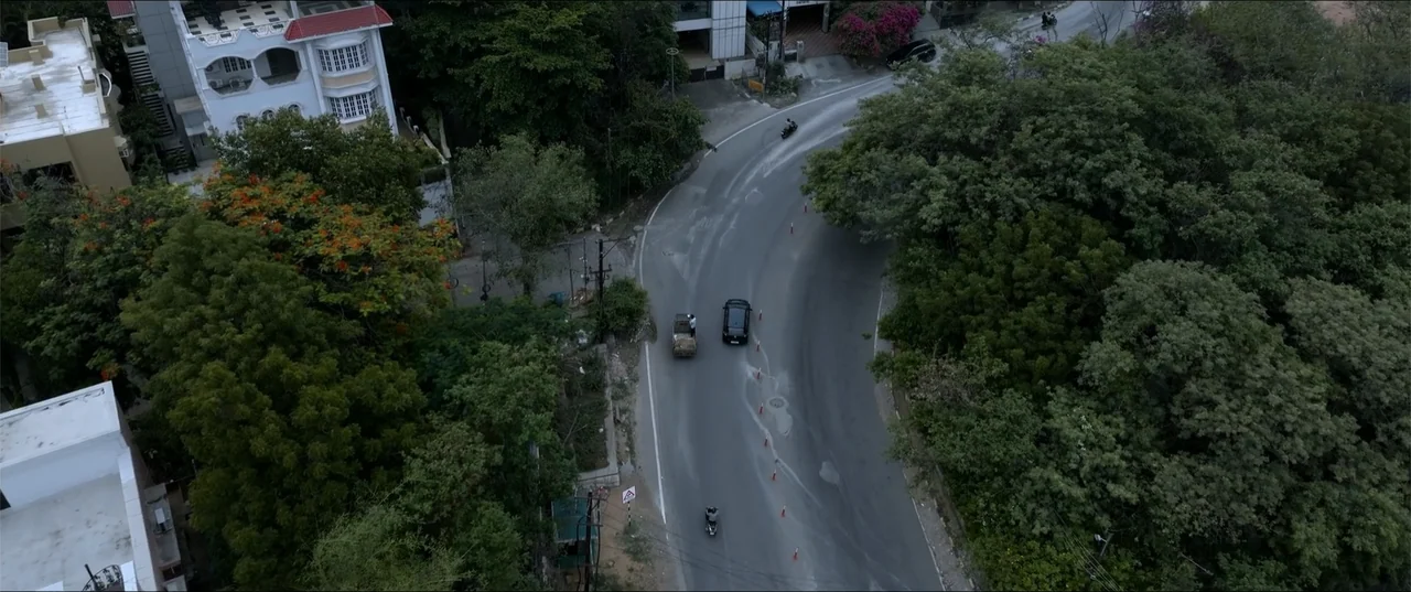 Cheekatilo (2026) movie still: wide — A high-angle view shows a winding asphalt road surrounded by dense green trees and vegetation. Two cars and a…