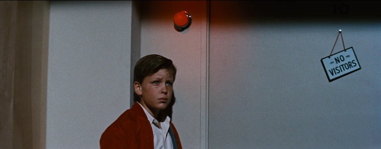 Bigger Than Life (1956) movie still: medium — A plain wall in a hallway with an orange warning light and a sign. A young boy stands by the wall…