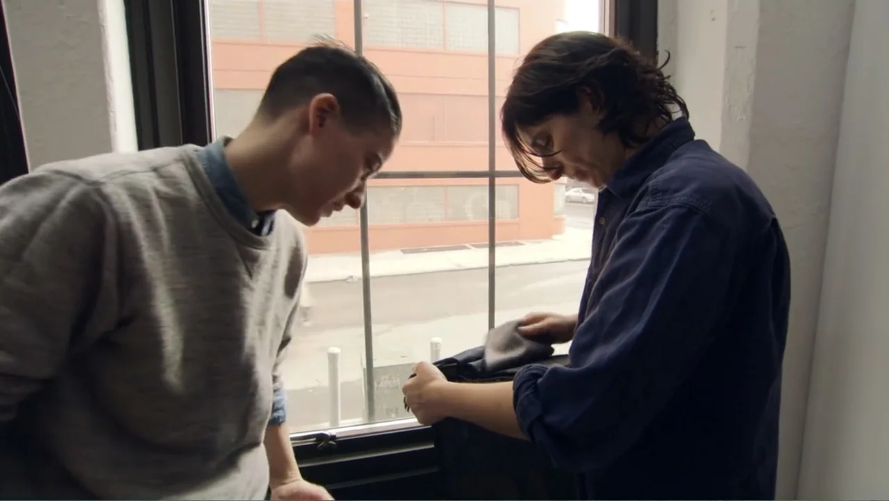 Suited (2016) movie still: medium — An interior office space with a large window looking out onto an urban street. Two men stand together by the…