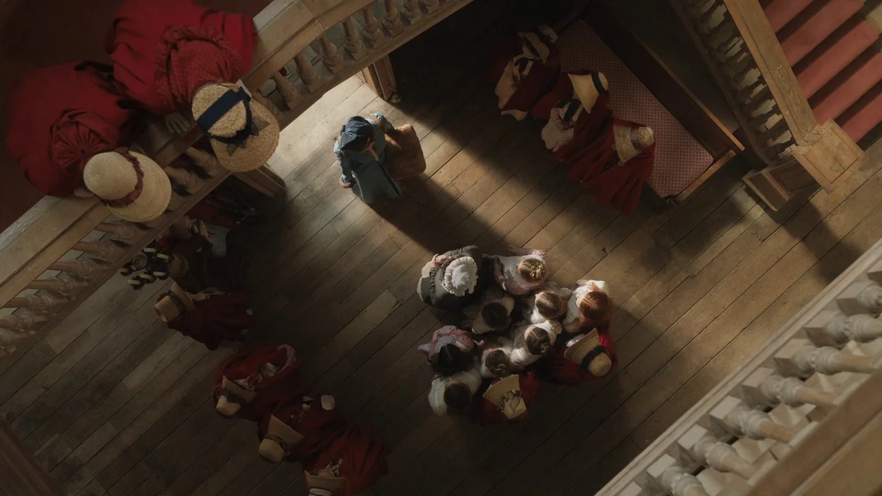 Emma. (2020) movie still: wide — An overhead view shows a wooden landing where a woman stands near a group of children. The staircase structure…