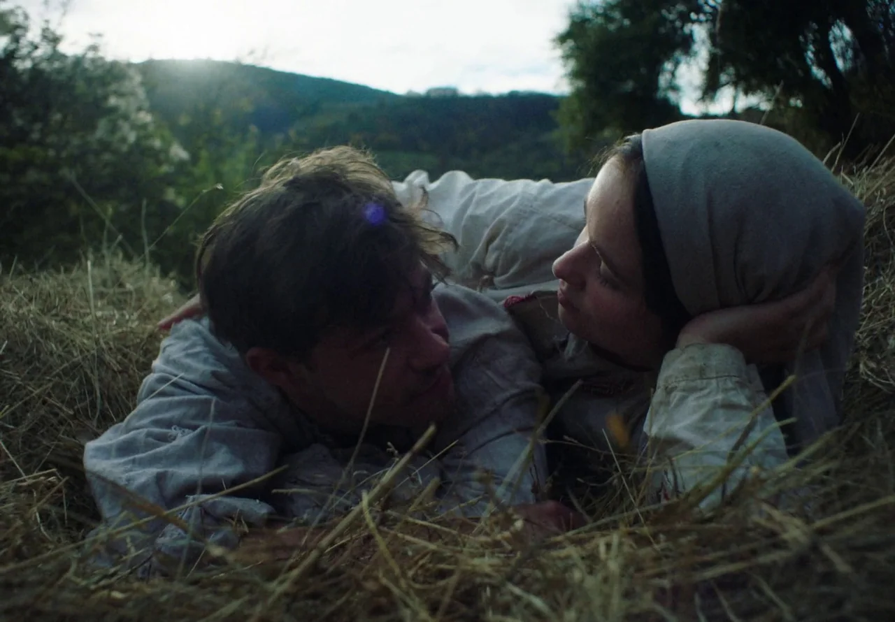 You Won’t Be Alone (2022) movie still: medium — A pair of people lie side by side in a field of harvested hay. They are looking directly at each…