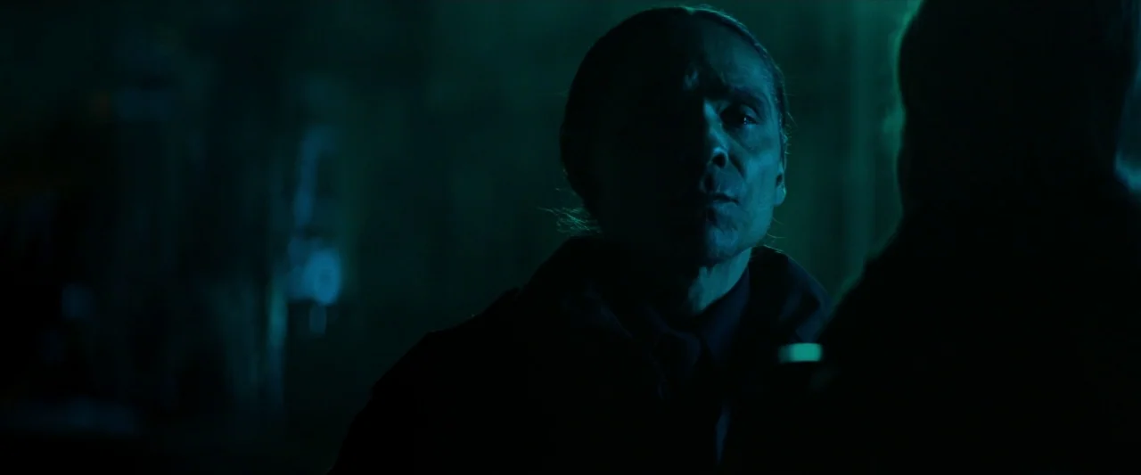 The Silencing (2020) movie still: closeup — A dimly lit room with surfaces mostly hidden in deep shadow. A man stands in the middle distance while…