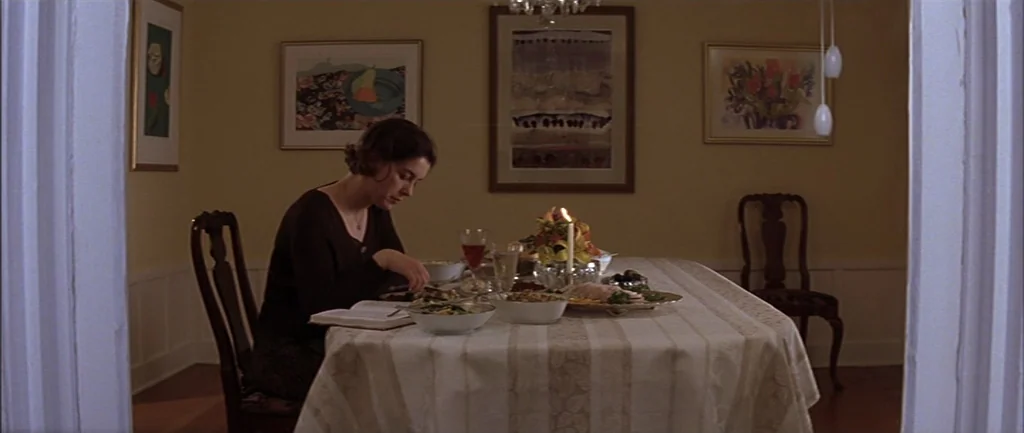 Rushmore (1998) movie still: medium — A quiet dining room with yellow walls and framed artwork above a wooden table. A woman sits alone at the table…