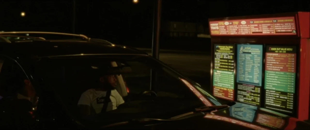 Sermon on the Mount (2019) movie still: medium — A man sits inside a dark car parked at a fast food drive-thru lane. He faces a large illuminated…