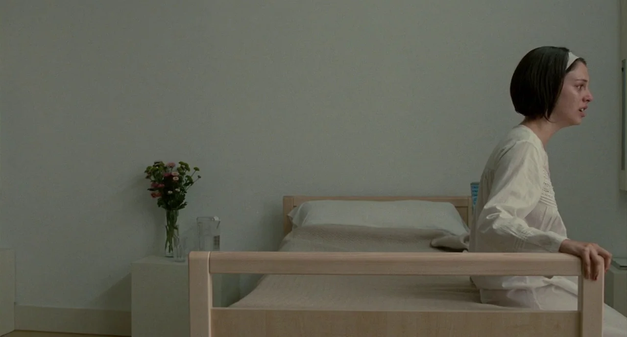The Skin I Live In (2011) movie still: medium — A minimalist white room contains a single light wood bed and a small nightstand. A woman in a white…