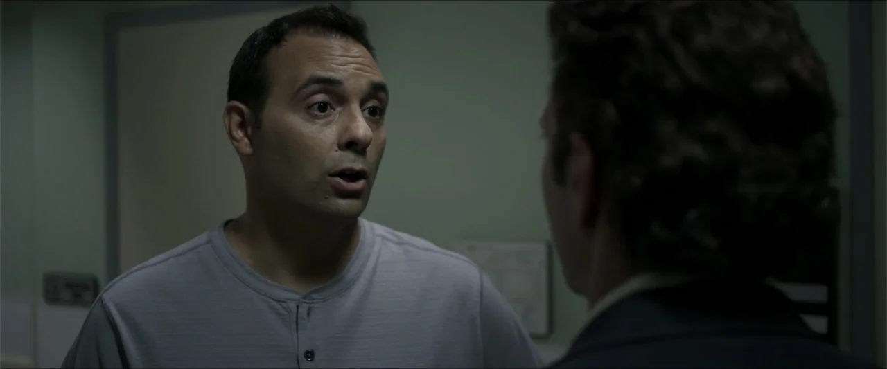 Heavenly Deposit (2019) movie still: medium — A dimly lit office space with plain grey walls and indistinct background features. A man in a grey…