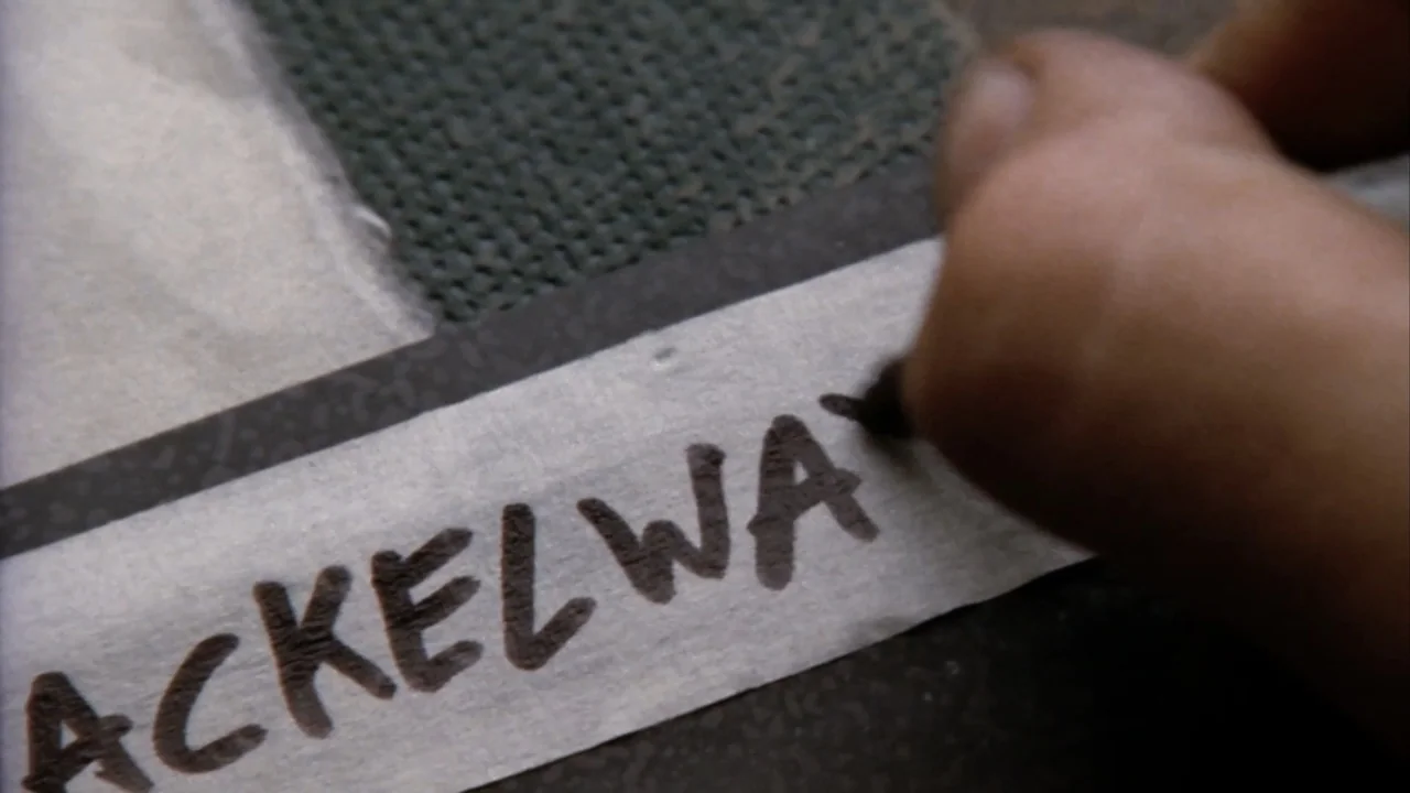 Suspect Zero (2004) movie still: closeup — A close-up view shows a hand using a black marker on a white strip. The label displays partial…