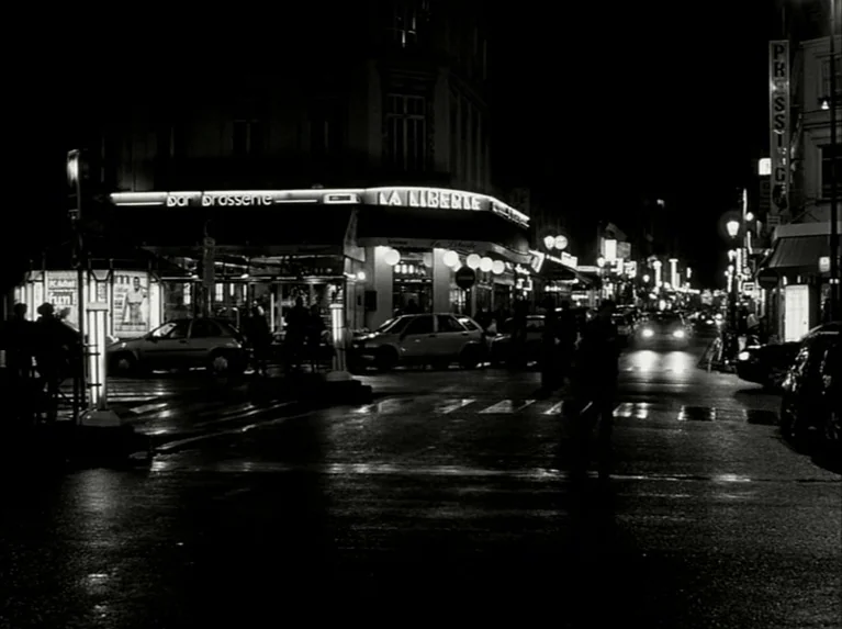 Eloge de L’amour (2001) movie still: establishing — A dark urban intersection at night features a glowing brasserie called La Liberte. Several…