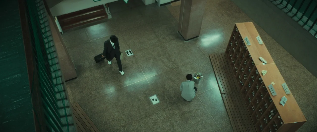 Monster (2023) movie still: wide — A school building entryway features a large wooden shoe rack and tiled floors. One person in a dark suit walks…